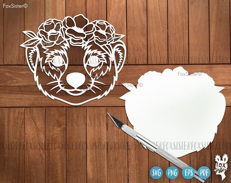 May include: White paper cut-out of a meerkat wearing a flower crown. The meerkat is facing forward with its eyes closed. The flower crown is made of simple flower shapes. The cut-out is on a wooden background.