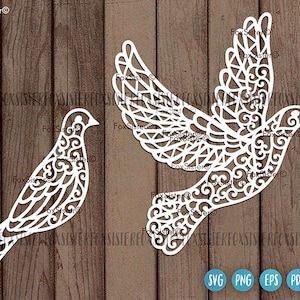 May include: Two white, intricate, laser-cut, paper doves with a filigree design. The doves are facing opposite directions. The larger dove has its wings spread out. The smaller dove is facing the left. The doves are on a wood background.