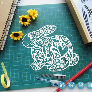 May include: White paper cut-out of a rabbit with a floral design and a heart. The rabbit is on a green cutting mat with a yellow tape measure, black and white patterned tape, and a blue and white patterned tape.