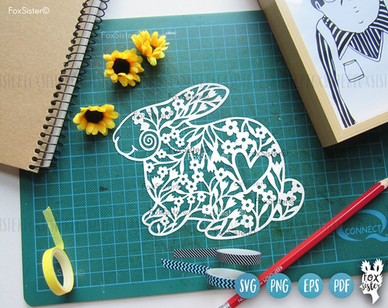 May include: White paper cut-out of a rabbit with a floral design and a heart. The rabbit is on a green cutting mat with a yellow tape measure, black and white patterned tape, and a blue and white patterned tape.