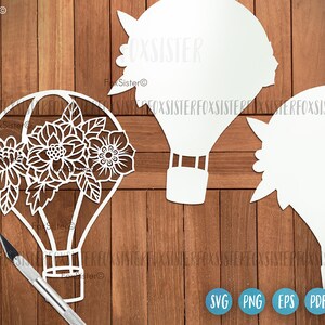 May include: White silhouette of a hot air balloon with floral designs, on a wood background. The balloon has a basket and a rope attached to it. The image also includes the text "FoxSister" and the file formats "SVG, PNG, EPS, PDF".