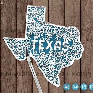 May include: A white paper cutout of the state of Texas, featuring intricate floral designs. The word "TEXAS" is cut out in white letters, set against a teal background. A craft knife rests on the wooden surface.