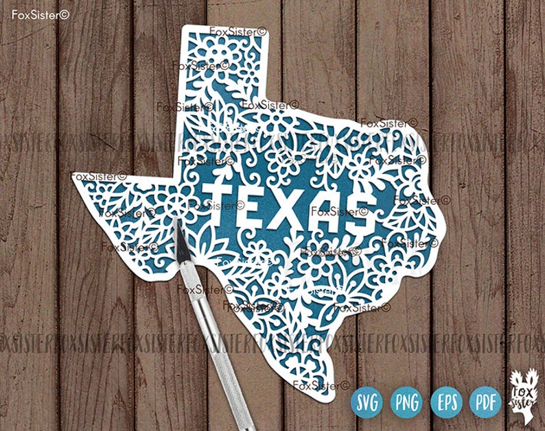 May include: A white paper cutout of the state of Texas, featuring intricate floral designs. The word "TEXAS" is cut out in white letters, set against a teal background. A craft knife rests on the wooden surface.