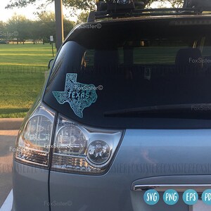 May include: A teal-colored Texas state outline sticker with the word "TEXAS" in the center, affixed to the rear window of a silver vehicle. The sticker has a detailed, patterned design. The image also shows the vehicle's taillight and wiper.