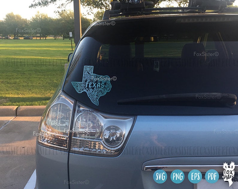 May include: A teal-colored Texas state outline sticker with the word "TEXAS" in the center, affixed to the rear window of a silver vehicle. The sticker has a detailed, patterned design. The image also shows the vehicle's taillight and wiper.