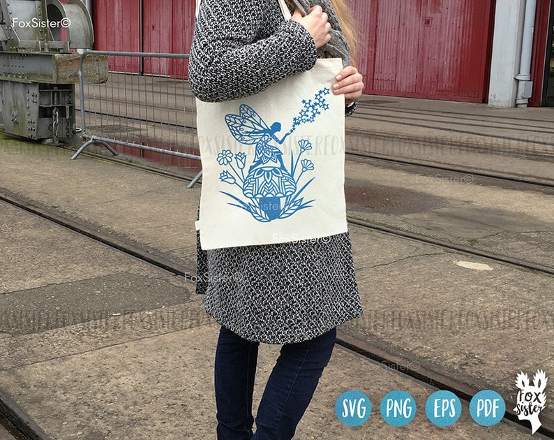May include: A white tote bag with a blue fairy design. The fairy is sitting on a mushroom and holding a wand with stars. The bag has the text "Fox Sister" on it.