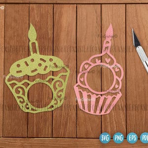 May include: Two decorative cupcake cutouts, one green and one pink, with candle toppers. The green cupcake has a dotted top and swirl details, while the pink one features heart accents. A craft knife rests beside them on a wooden surface.