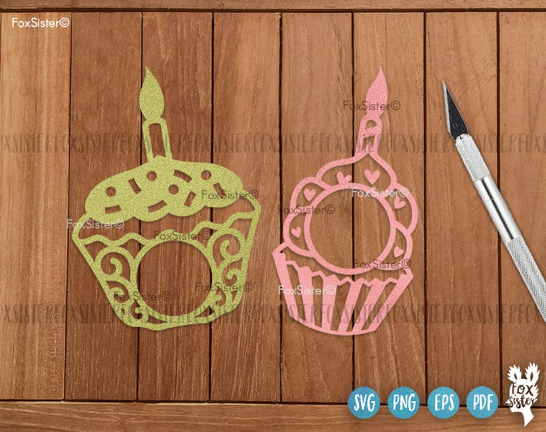 May include: Two decorative cupcake cutouts, one green and one pink, with candle toppers. The green cupcake has a dotted top and swirl details, while the pink one features heart accents. A craft knife rests beside them on a wooden surface.