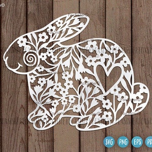 May include: White paper cut-out of a rabbit with a floral design and a heart in the center. The rabbit is surrounded by a wood grain background.