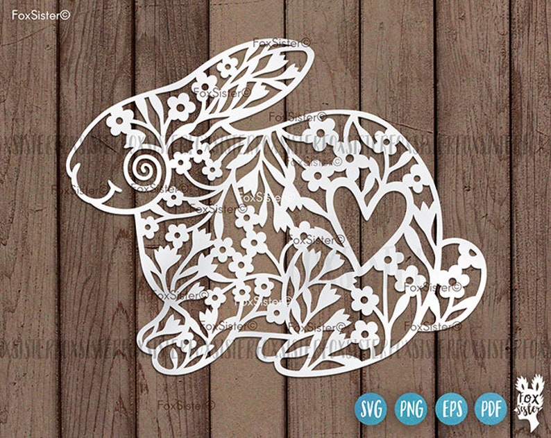 May include: White paper cut-out of a rabbit with a floral design and a heart in the center. The rabbit is surrounded by a wood grain background.