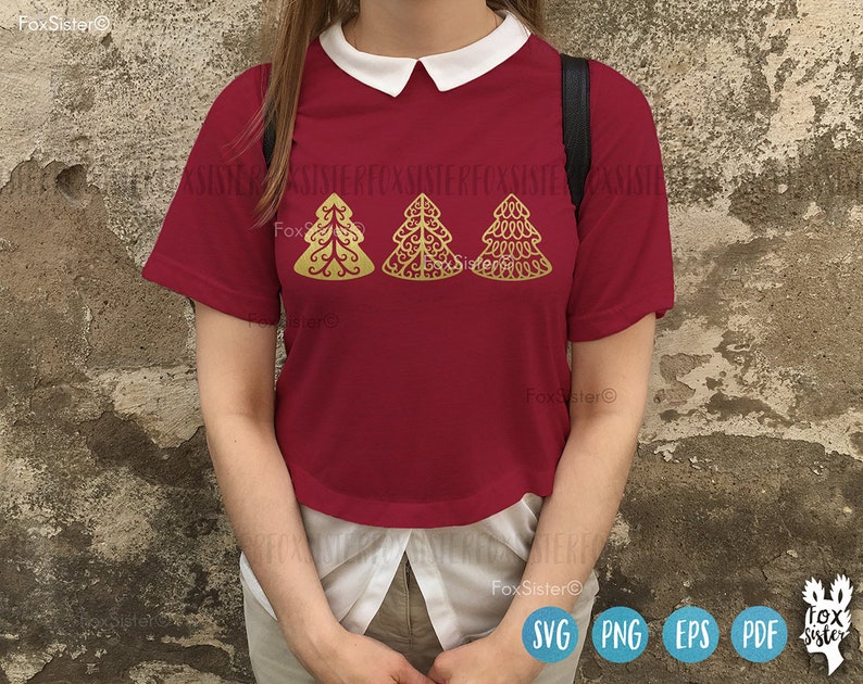 May include: A red short-sleeved shirt with a white collar features three gold Christmas tree designs. The trees are stylized and have a decorative, intricate pattern. The shirt is worn by a person standing in front of a textured wall.