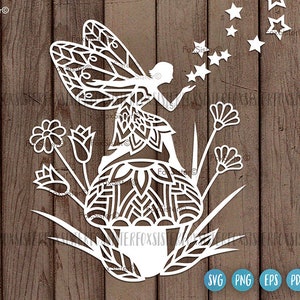 May include: White paper cut-out design of a fairy with wings, sitting on a mushroom with flowers and stars around her. The design is on a wood background.