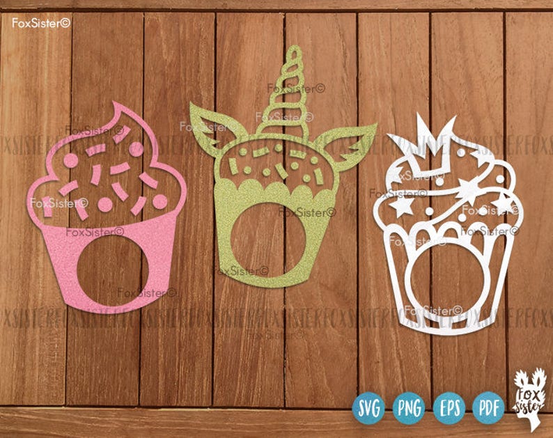 May include: Three glittery cupcake-shaped cutouts in pink, green, and white. The pink cupcake has sprinkles, the green one has a unicorn horn, and the white one has a crown. Each has a circular opening. The FoxSister logo is visible.