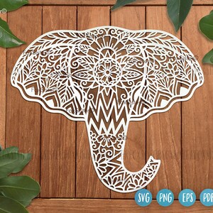 May include: White, intricately cut elephant design with floral and geometric patterns. The elephant is set against a wooden background with green leaves. The image includes SVG, PNG, EPS, and PDF file type icons.
