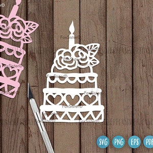 May include: White paper cut-out of a cake with a single candle and a rose on top. The cake is decorated with hearts and a scalloped edge. The cake is on a wooden background.