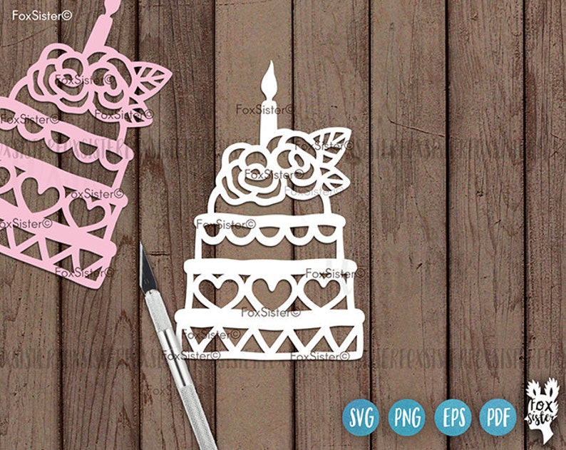 May include: White paper cut-out of a cake with a single candle and a rose on top. The cake is decorated with hearts and a scalloped edge. The cake is on a wooden background.