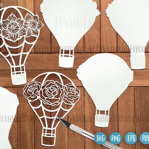 May include: A set of four white paper cut-out hot air balloons with floral designs. The balloons are in various shapes and sizes, with some featuring a single large flower and others with multiple smaller flowers. The balloons are arranged on a wooden surface.