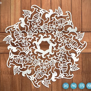 Puede incluir: Diseño de mandala blanco intrincado con patrones florales y de ondas. El diseño está cortado en papel y está sobre un fondo de madera.