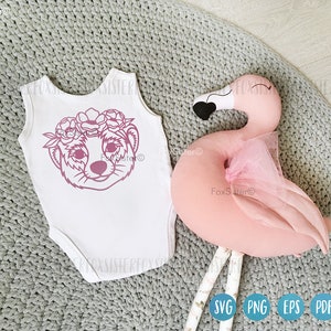 May include: A white onesie with a pink outline of a meerkat wearing a flower crown. The onesie is on a gray knitted surface next to a pink flamingo pillow.