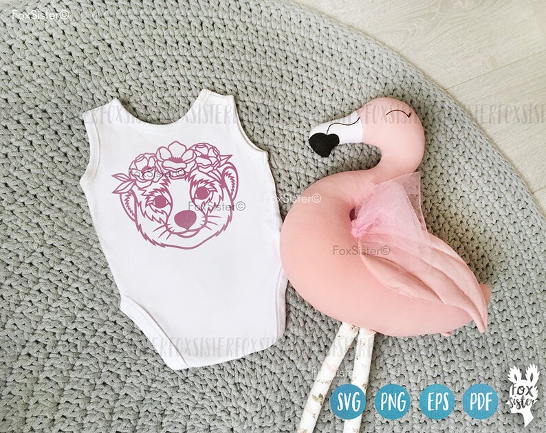May include: A white onesie with a pink outline of a meerkat wearing a flower crown. The onesie is on a gray knitted surface next to a pink flamingo pillow.