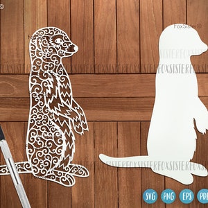 May include: A white silhouette and a detailed, intricate cutout of a meerkat standing on its hind legs with its arms raised. The meerkat is facing to the right. The background is a wood grain pattern.