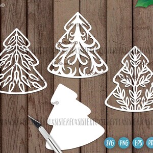 May include: Four white paper cut-out Christmas tree designs. The trees are different styles, with one being a simple outline, one with a leafy design, one with a swirling design, and one with a more intricate design. The trees are on a wood background.
