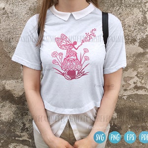 May include: A white t-shirt with a pink silhouette of a fairy blowing dandelion seeds. The fairy is sitting on a mushroom with a mandala design. The design is surrounded by pink flowers.