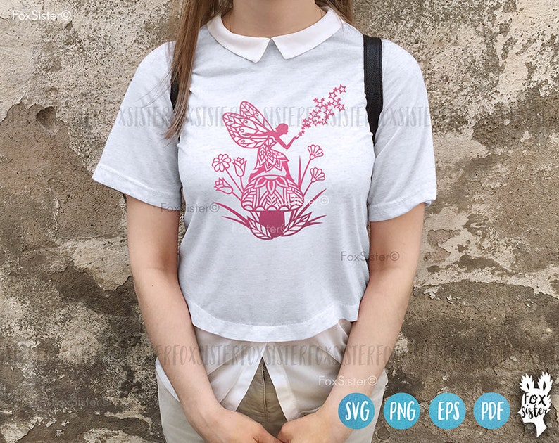 May include: A white t-shirt with a pink silhouette of a fairy blowing dandelion seeds. The fairy is sitting on a mushroom with a mandala design. The design is surrounded by pink flowers.