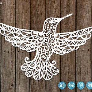 Könnte beinhalten: Weiße, komplizierte Silhouette eines Kolibris mit Mandala-Muster. Der Kolibri ist nach rechts gerichtet und hat seine Flügel ausgebreitet. Der Hintergrund ist eine Holzmaserung.