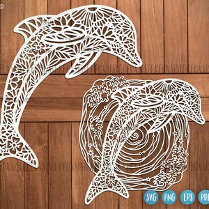 Könnte beinhalten: Zwei weiße, lasergeschnittene Delfin-Designs. Ein Delfin ist eine freistehende Silhouette, der andere befindet sich in einem kreisförmigen Rahmen mit Wellen und Sternen. Beide weisen komplizierte Blumen- und Blättermuster auf. Der Hintergrund ist Holzmaserung.