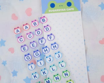 May include: A sheet of alphabet stickers with frog faces in various colors. The letters A through S are visible. A notepad with the text "SUGARSTAR CAFE" is also in the image. The background is white with pink hearts and blue stars.