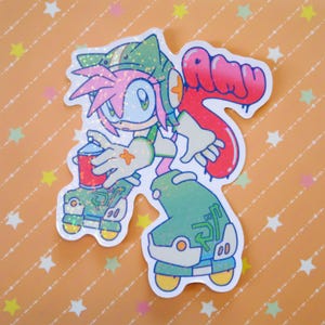 Könnte beinhalten: Ein holografischer Aufkleber mit einer Zeichentrickfigur mit rosa Haaren, grüner und weißer Kleidung und einer roten Sprühdose. Die Figur steht auf Rollschuhen und hält die Dose. Das Wort "AMY" ist in roter Graffiti-Schrift geschrieben.