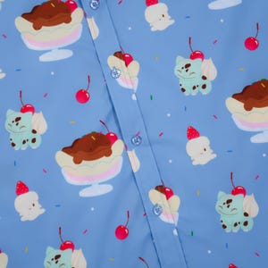 Puede incluir: Una camisa de botones azul claro con un patrón repetitivo de criaturas de dibujos animados, banana splits y helados cubiertos con cerezas. La tela está decorada con pequeñas chispas de colores y estrellas blancas por todo el diseño.