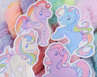 May include: Four colorful stickers of cartoon ponies with glitter accents. Each pony has a unique color scheme, including pink, blue, purple, and white, with rainbow manes and tails. The stickers feature bows and are set against a backdrop of pastel yarn.