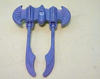 May include: A purple plastic toy weapon with a bat-like design. The toy has two curved blades extending from a central body with a circular detail. The color is a muted purple.