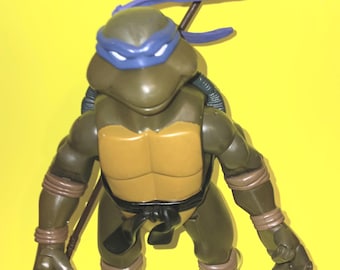 May include: A dark olive green action figure of a turtle character wearing a blue eye mask and a blue headband. The figure has a tan chest plate, a black belt, and brown wrist and ankle wraps. A long brown staff is positioned behind the figure's back.
