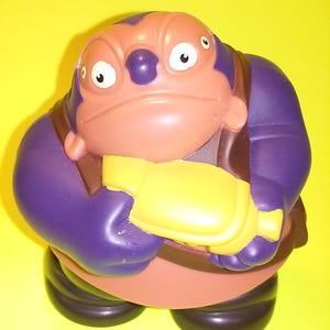 Op de afbeelding: Een paars en bruin plastic speelgoedfiguur met drie ogen en een geel pistool. Het figuur draagt een paarse jas en een zwarte broek.