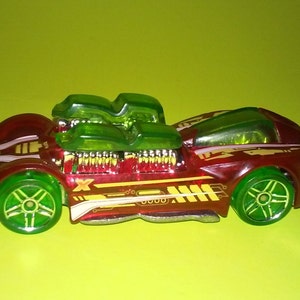 May include: A red toy car with green transparent engine details and yellow accents. The car has white stripes and a black "X" on the side.