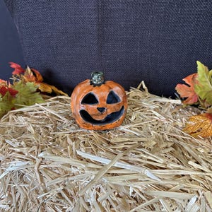 May include: A small, orange ceramic jack-o'-lantern with a smiling face and a green stem. It sits on a bed of straw, surrounded by colorful autumn leaves. The Halloween-themed decoration is perfect for fall.