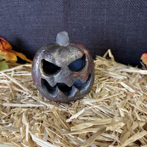 May include: A metallic-colored, carved pumpkin with a menacing face. The pumpkin sits on a bed of straw, with fall leaves in the background. The pumpkin's eyes and mouth are cut out, revealing a dark interior, perfect for Halloween decor.