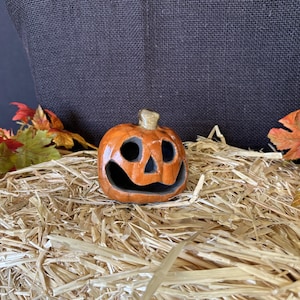 May include: A small, orange ceramic jack-o'-lantern with carved eyes and a mouth, set on a bed of straw. The pumpkin has a stem and is surrounded by colorful autumn leaves, creating a festive Halloween scene.