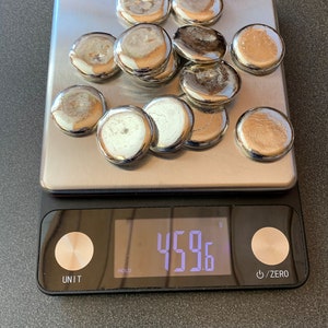 May include: A collection of shiny, silver-colored, round objects are arranged on a digital scale. The scale displays a weight of 459.6 grams. The objects have a reflective surface.