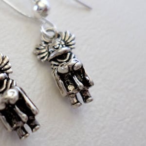 May include: A pair of silver earrings featuring a small, detailed charm of a figure wearing a hat and holding a staff. The earrings have a simple, silver hook design.