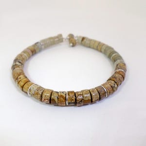 Puede incluir: Un brazalete de cuentas cilíndricas en tonos tierra, con espaciadores plateados. Las cuentas tienen un aspecto natural y texturizado, con variaciones de color que van del beige al marrón y al gris. El brazalete tiene forma circular.