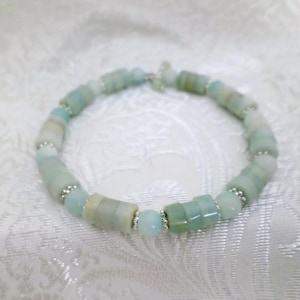 Puede incluir: Una pulsera de cuentas con cuentas cilíndricas de color azul claro y verde, intercaladas con cuentas más pequeñas de color plateado. La pulsera está hecha de piedras naturales y se muestra sobre una superficie blanca texturizada.