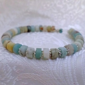 Puede incluir: Una pulsera hecha a mano con una variedad de cuentas cilíndricas de colores en tonos de azul, verde, amarillo y blanco. Las cuentas están ensartadas con detalles plateados, creando un accesorio único y elegante.