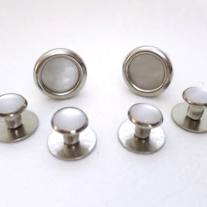 May include: A set of six silver-toned shirt studs. Two larger studs have a flat, circular base and a domed, white center. Four smaller studs have a flat base and a domed, silver-toned top.