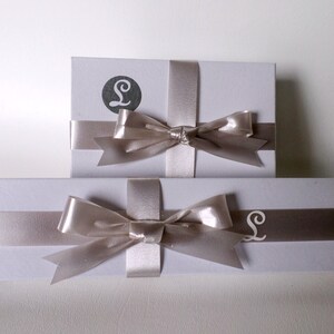 May include: Two white gift boxes with a silver ribbon and bow. The top box has a silver letter "L" on the front. The bottom box has a silver letter "L" on the front.