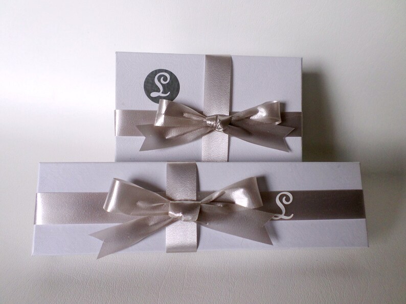 May include: Two white gift boxes with a silver ribbon and bow. The top box has a silver letter "L" on the front. The bottom box has a silver letter "L" on the front.