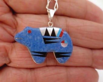 May include: A blue bear pendant with inlaid details. The bear has a red eye, black stripes, and a silver bail. The pendant is on a silver chain. The bear pendant is a unique piece of jewelry.